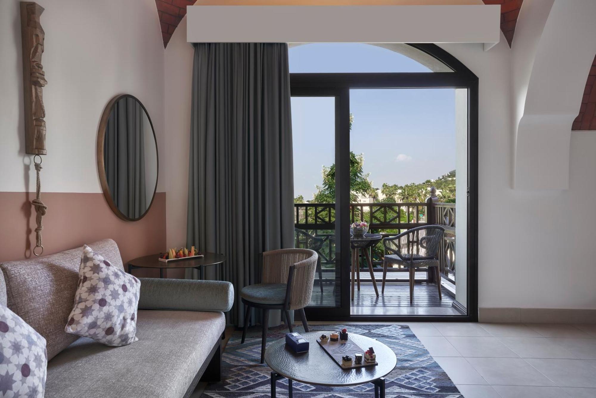 The Cove Rotana Resort - Ras Al Khaimah Kültér fotó The photo shows a modern and stylish hotel room. There is a comfortable gray sofa with decorative pillows, a round coffee table in front of it, and a couple of chairs nearby. Large curtains frame a glass door that leads to a balcony, providing natura
